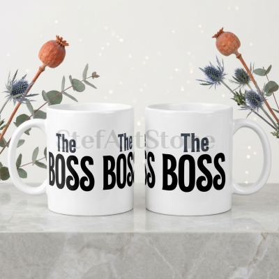 Две бели керамични чаши с черно-сив надпис The Boss, показани върху светъл фон с цветя.