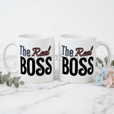 Две бели керамични чаши с надпис The Real Boss, с тъмна типография и розовочервен акцент върху думата Real.